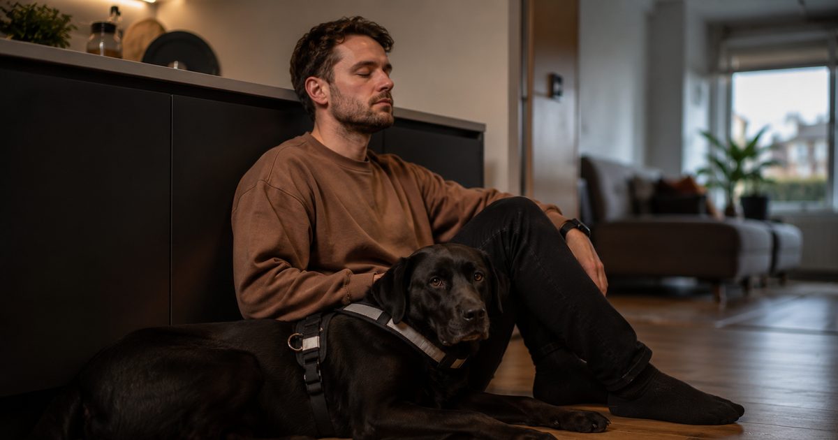 A man sits on a wooden floor leaning against a sofa with a black Labrador assistance dog lying beside him in a harness